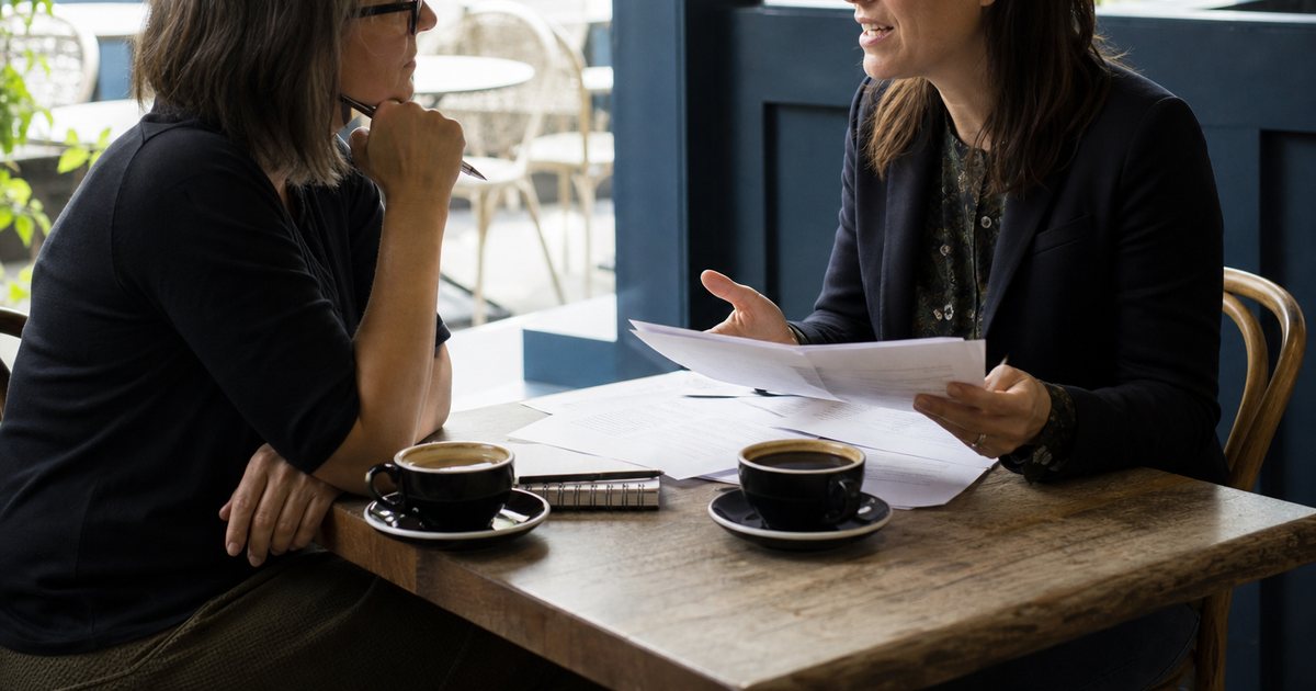 Nonprofit development director and a freelance writer meeting at a cafe table with printed proposal drafts, notebooks, and two cups of coffee