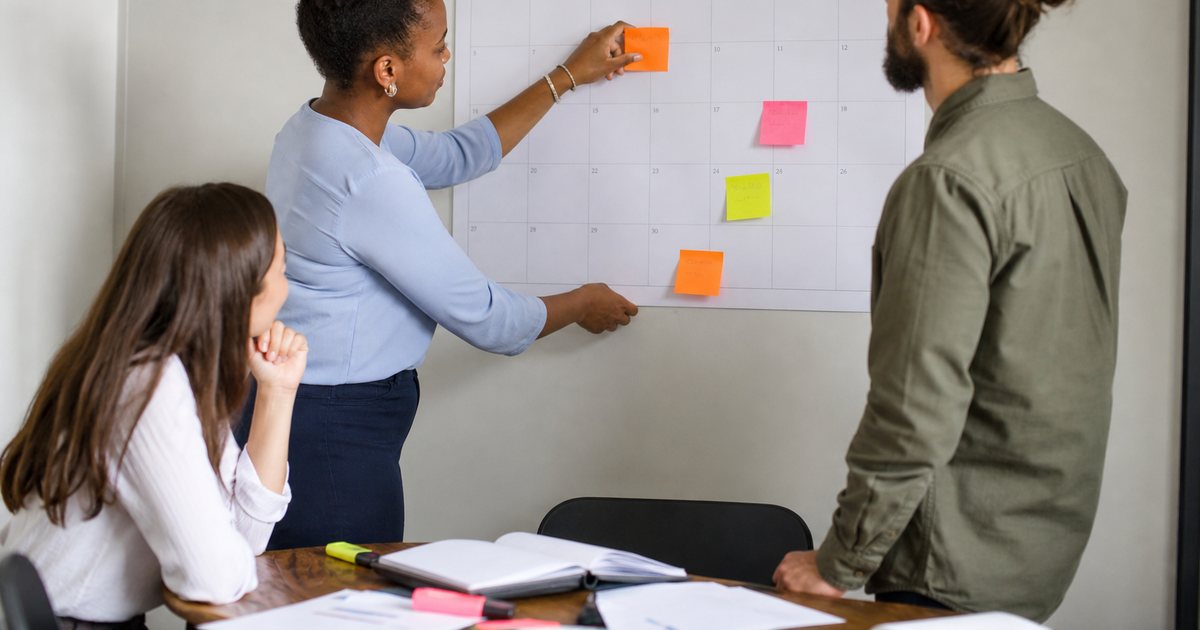 Small nonprofit team of three gathered around a wall calendar in a bright office, one person placing a sticky note on a date, papers and highlighters on the table